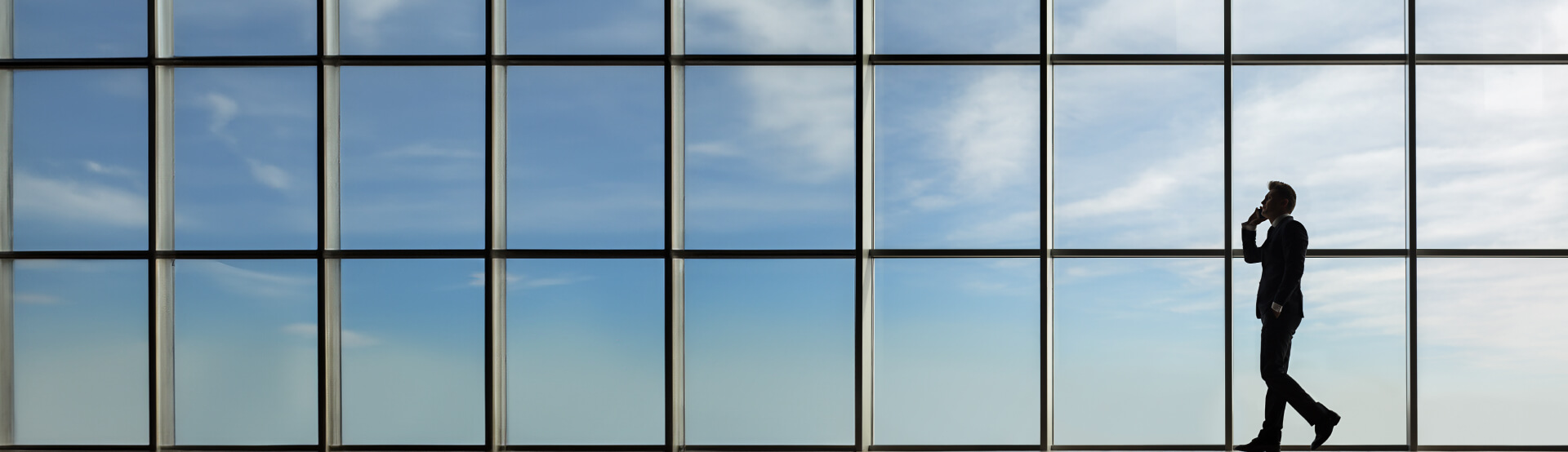 A man on the phone walking from right to left, with windows showing the sky in the background